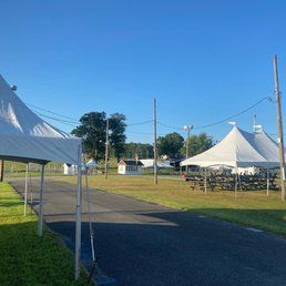 An asphalt road leads to white tents set up on a grassy area under a clear blue sky. Utility poles are visible.