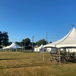 Several white tents are set up on a grassy field under a clear blue sky. Picnic tables are in the foreground.
