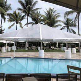 A clear tent set up by a pool. Palm trees are visible in the background under a cloudy sky.