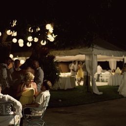 Nighttime outdoor party with a tent, string lights, and people mingling. Guests seated at tables.