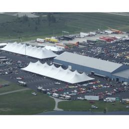 Aerial view of an outdoor event with large white tents, a large building, and a crowded parking area filled with vehicles.