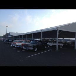Classic cars parked under a long white tent structure on a paved lot. Blue sky overhead.
