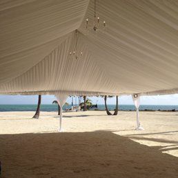 Tent on a beach, with ocean visible. White fabric roof and support posts, tan sand, blue water, and palm trees.