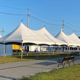Several white tents set up outdoors on a paved area with benches, grass, and utility poles, under a blue sky.