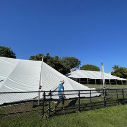 A person walks past a white tent with a sloped roof, under a blue sky. Another tent is visible in the distance.
