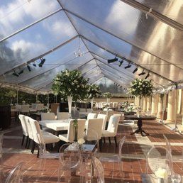 A clear event tent with tables and white chairs set for a gathering. Floral arrangements are on the tables.