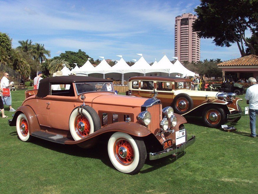 Two vintage cars on display at an outdoor event, one brown convertible and one woody-style car, with tents and a building in the background.