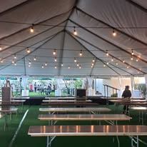 Inside a large white tent, long tables and benches are set up. String lights hang overhead, and people are visible in the background.
