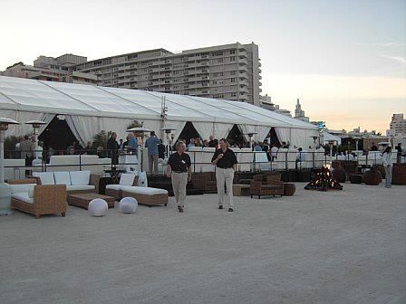 Two men walk past outdoor lounge furniture in front of a large white tent, with a high-rise building in the background.