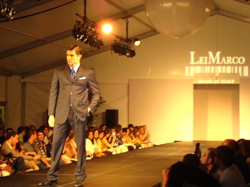 A model in a dark suit poses on a runway during a fashion show, under bright lights, with an audience watching.