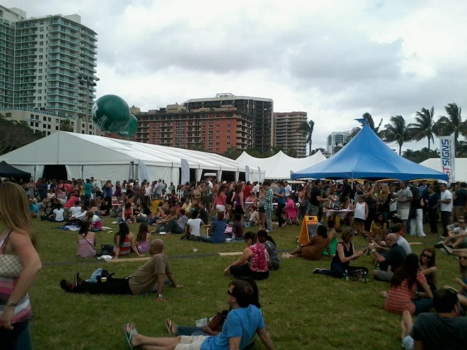Large outdoor festival with many people on a grassy field; white tents, a blue tent, and skyscrapers in the background.