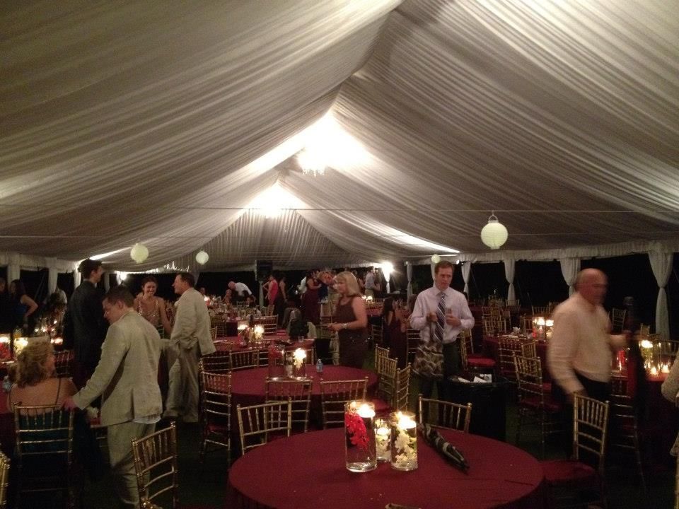 Inside a large white tent, people mingle at tables set with red tablecloths and candles.