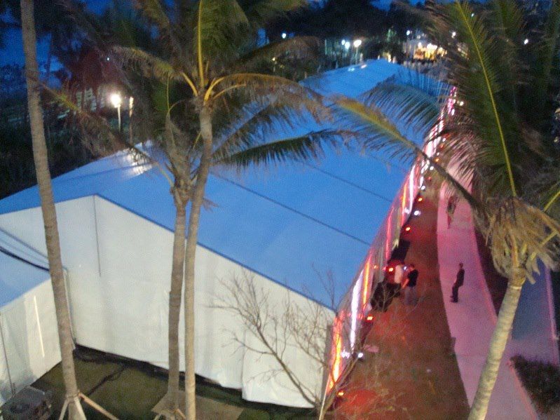 A long white event tent at night, with a blue roof and palm trees in the foreground. People are gathered inside.