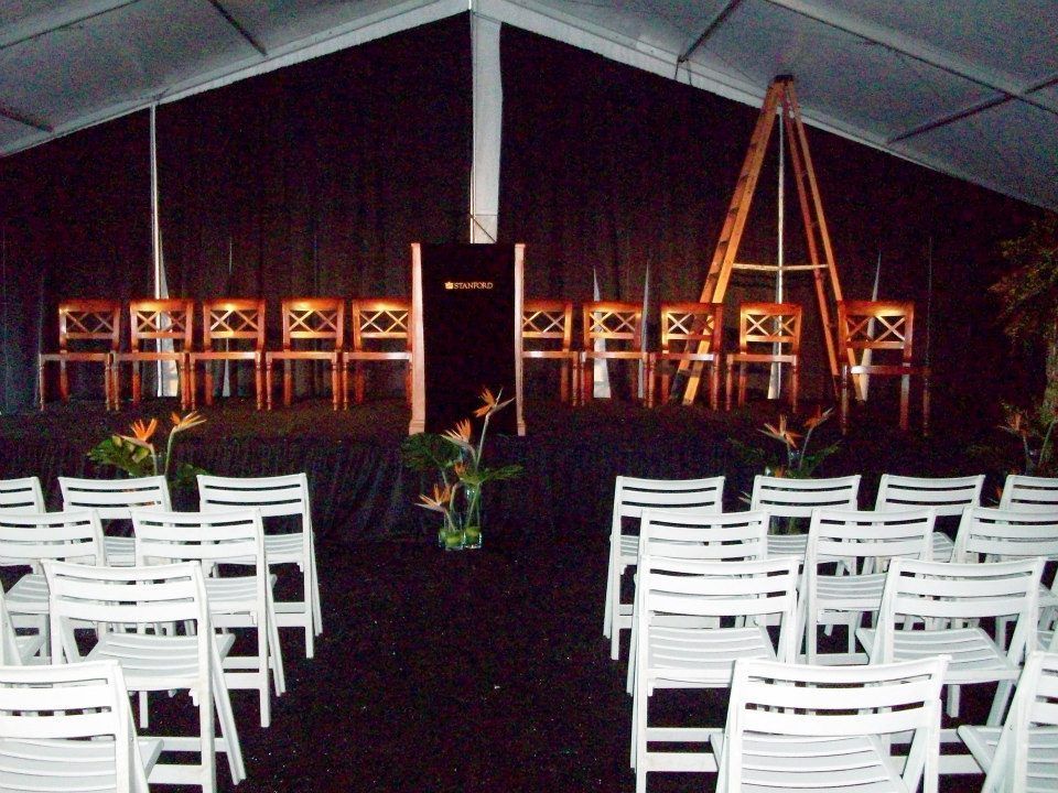 Rows of white chairs face a stage with dark backdrop, wooden chairs, and a podium under a tent.