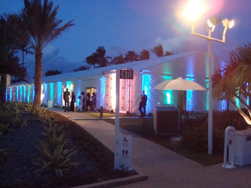 An outdoor event venue at night with colorful lights illuminating the building. People stand near the entrance with palm trees and a pathway in the foreground.