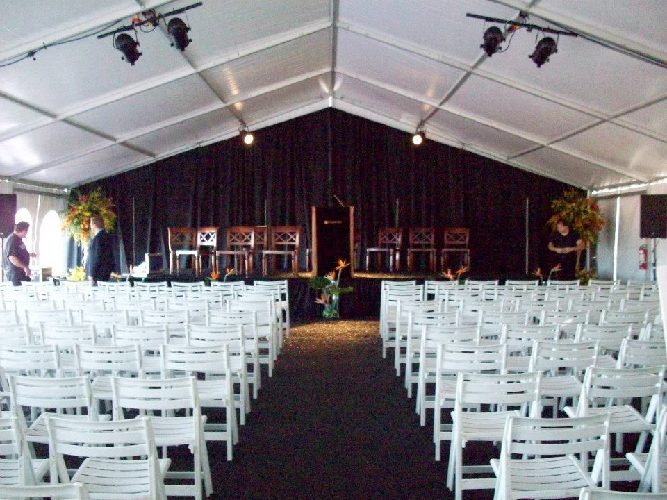 Inside a white tent set up for an event. Rows of white chairs face a stage with a black curtain and podium.