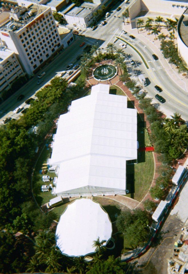 Aerial view of a large white tent in a park surrounded by greenery and buildings. A fountain is visible near a curved road.