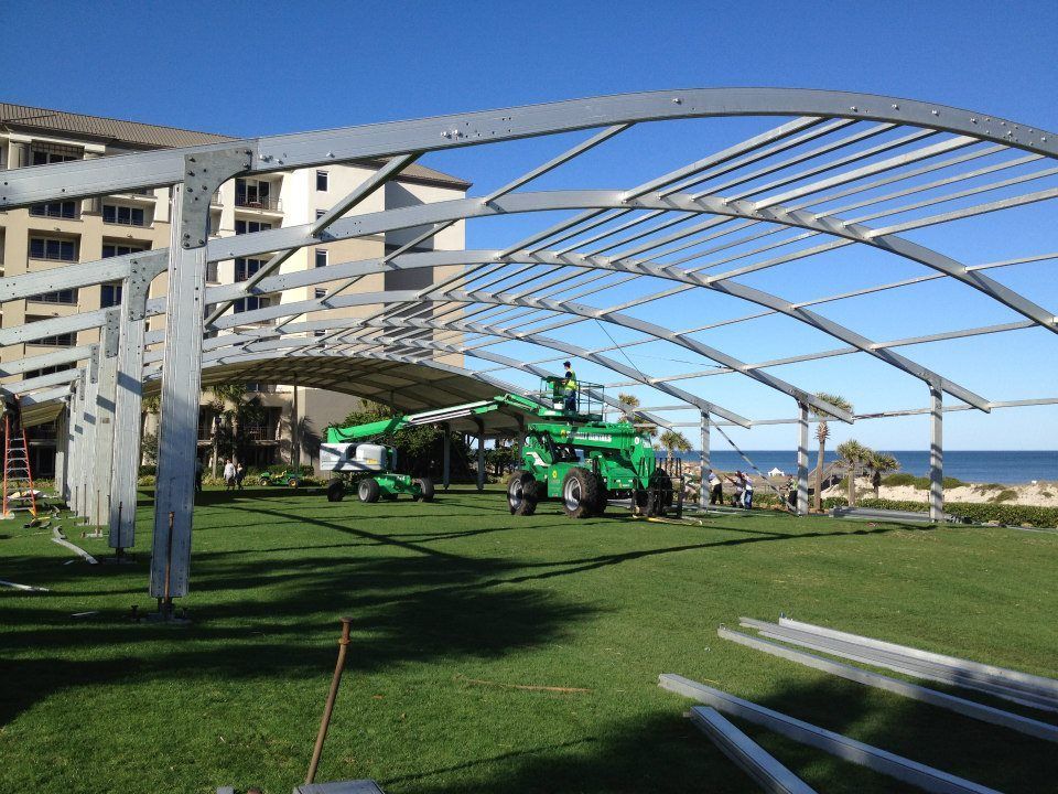 A metal tent frame being assembled on a grassy lawn near a beach. Workers use machinery to connect the beams.
