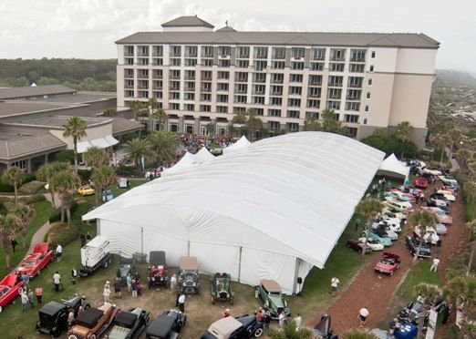 An aerial view of a car show with classic cars on display near a large white tent and a multi-story hotel.
