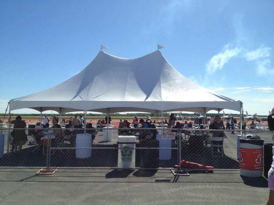 Large white tent set up on an airfield. People are gathered inside and around the tent, likely for an outdoor event under a bright blue sky.