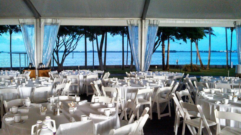 A large, white-themed event tent overlooking a body of water. Round tables with white tablecloths and chairs fill the space.
