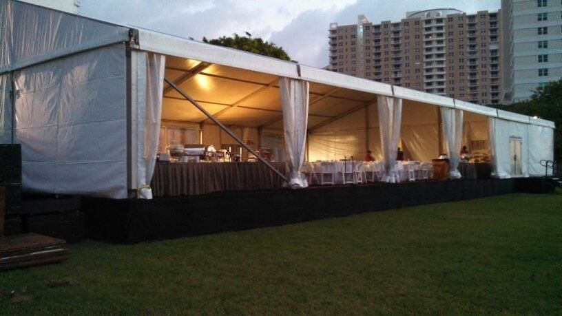 Long white tent set up outdoors, likely for an event. Tables with white chairs are visible inside, with a tall building in the background.