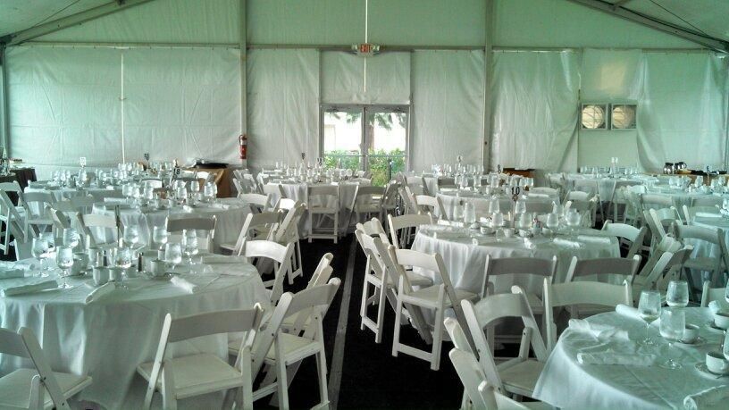 White tables and chairs set for a formal event inside a large tent. Tables are set with glassware and plates.