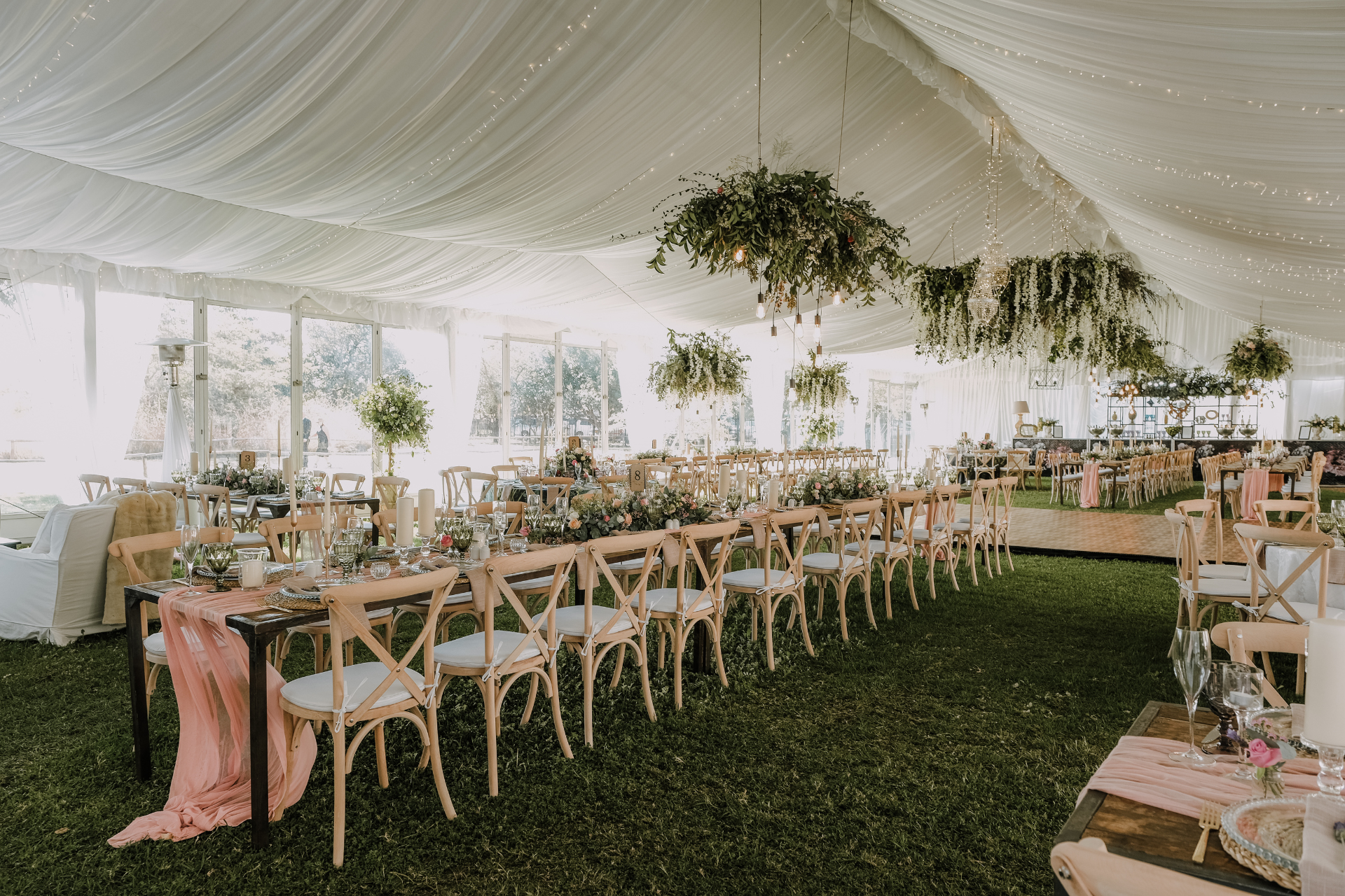 Wedding reception in a white tent with long tables, wooden chairs, and floral decorations.