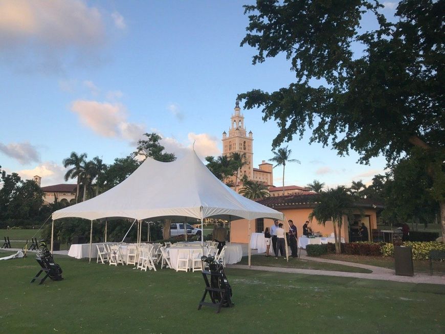 A white tent set up on a green lawn, with a tall building in the background under a blue sky. People stand nearby.