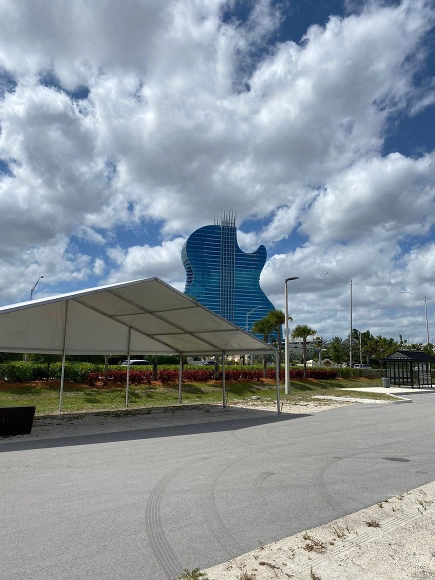 A large blue guitar-shaped hotel against a cloudy sky, with a white tent and paved area in the foreground.