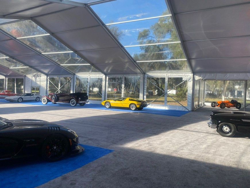 Classic cars on display under a large clear tent on a sunny day. Various colors, including black, yellow, and white.