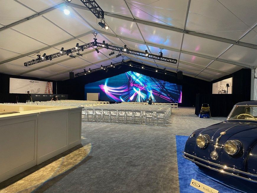 A large tent interior setup for an event. A blue classic car sits on a blue rug. A large digital screen displays colorful graphics.