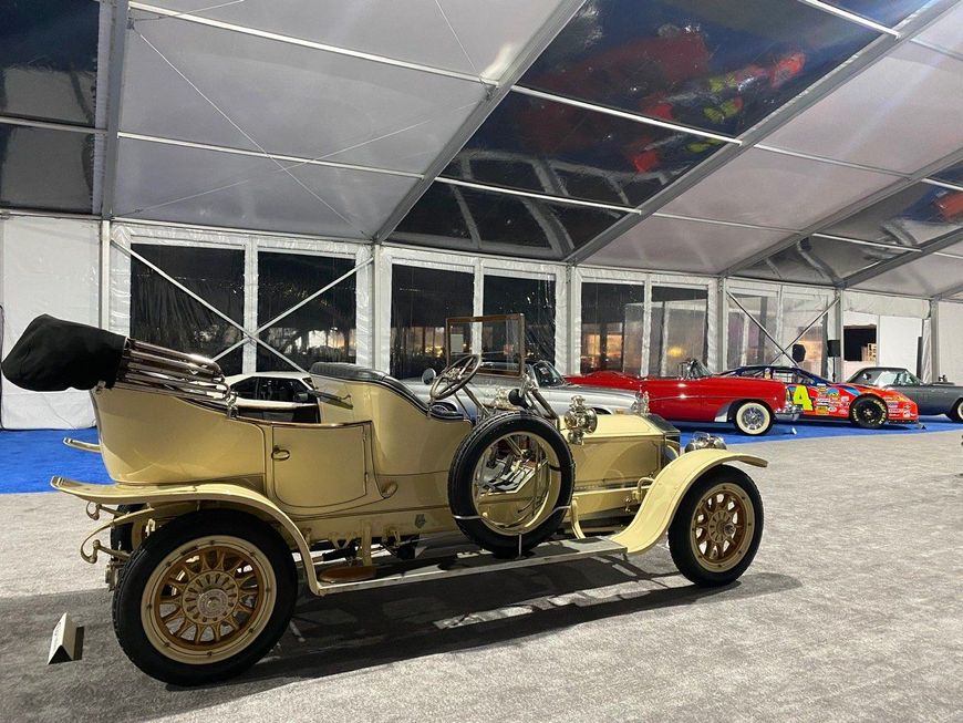 A vintage cream-colored car with gold wheels is displayed under a white tent. Other cars are visible in the background.