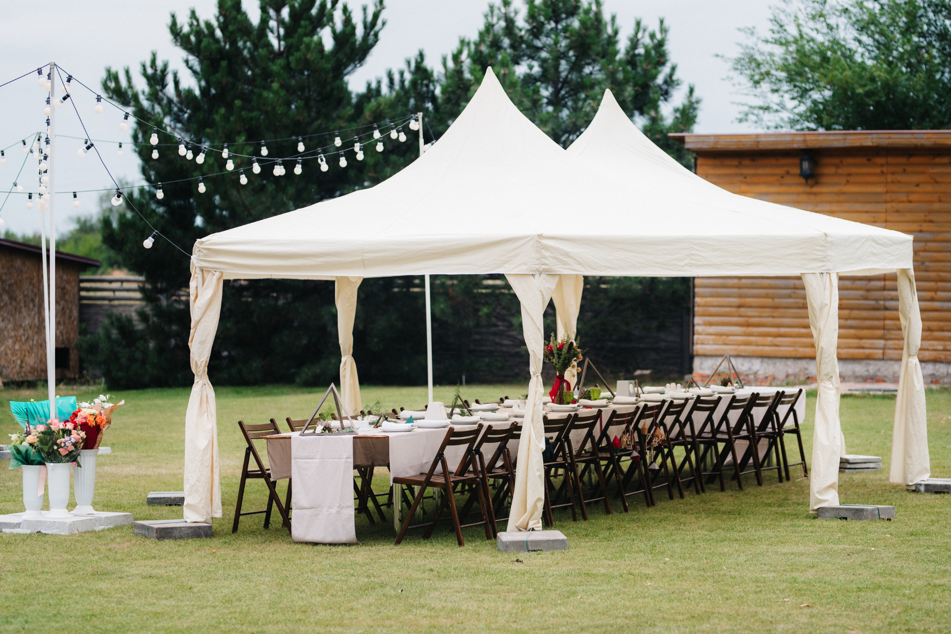 A long table set for a party under a white tent in a grassy yard, with string lights and colorful flowers.