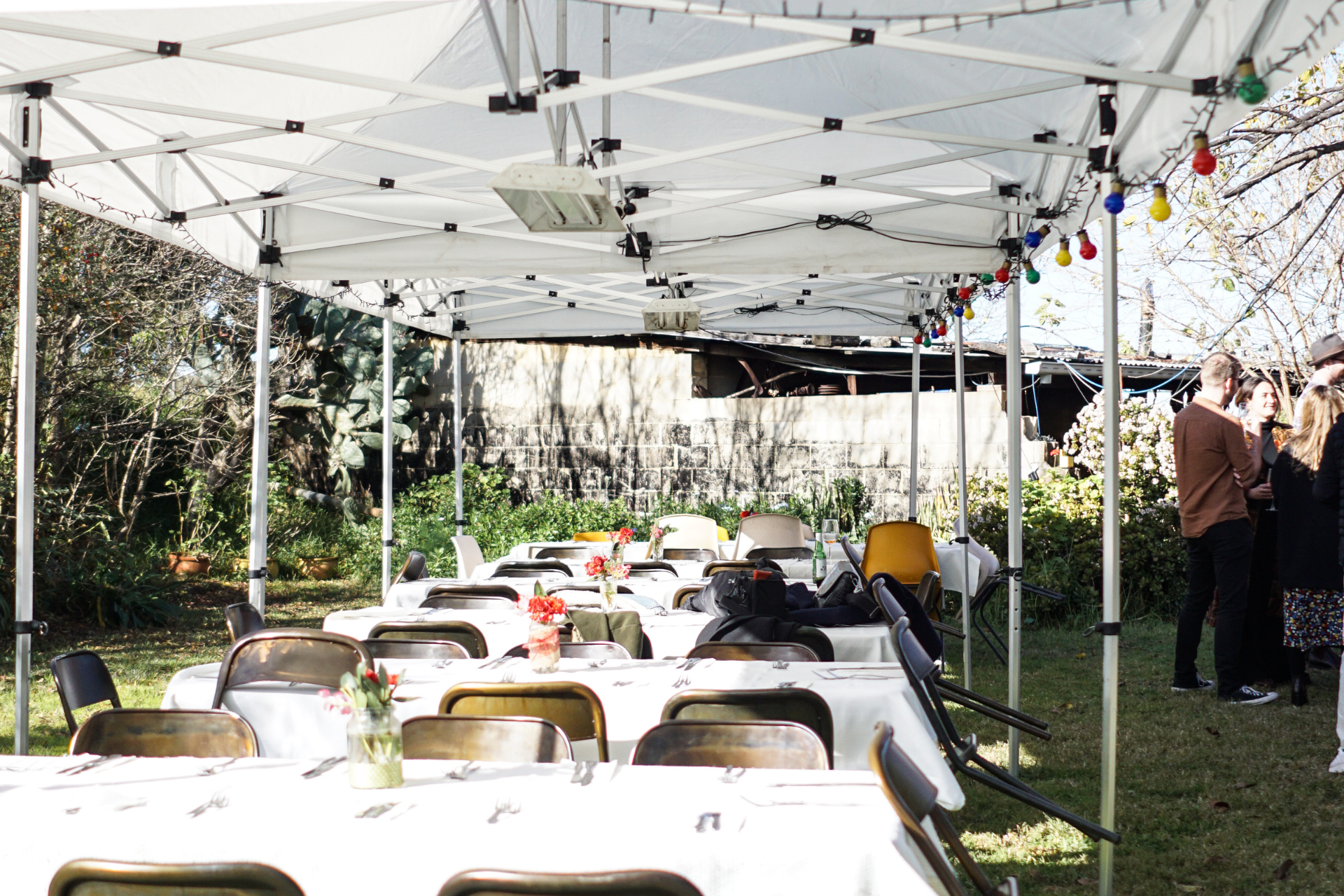 Outdoor party setup with long tables covered in white tablecloths, under a white canopy, strung with colorful lights. People stand near the tables in a grassy backyard.