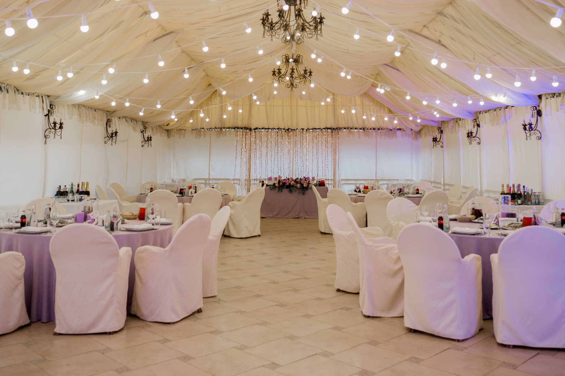 Inside a decorated tent, round tables set for a formal event, with white chair covers and string lights.