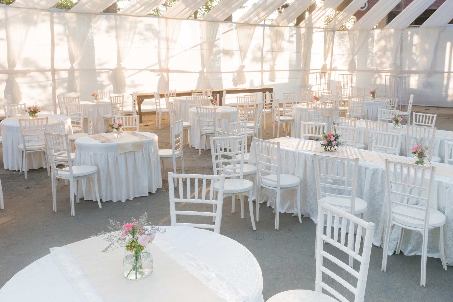 White-themed outdoor event space with round and rectangular tables, all covered in white linens. White chairs are arranged around the tables, set up for a gathering.
