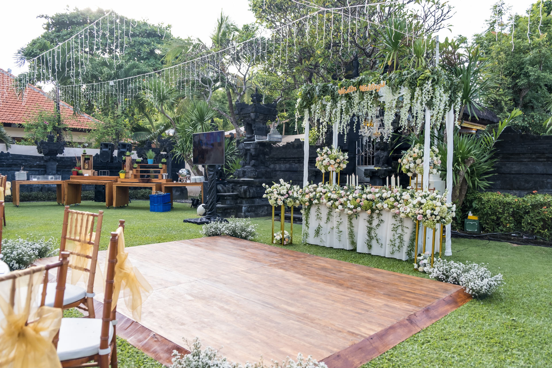 Wedding reception setup in a lush green garden. Wooden dance floor, decorated arch with white flowers, and chairs with gold accents are visible.