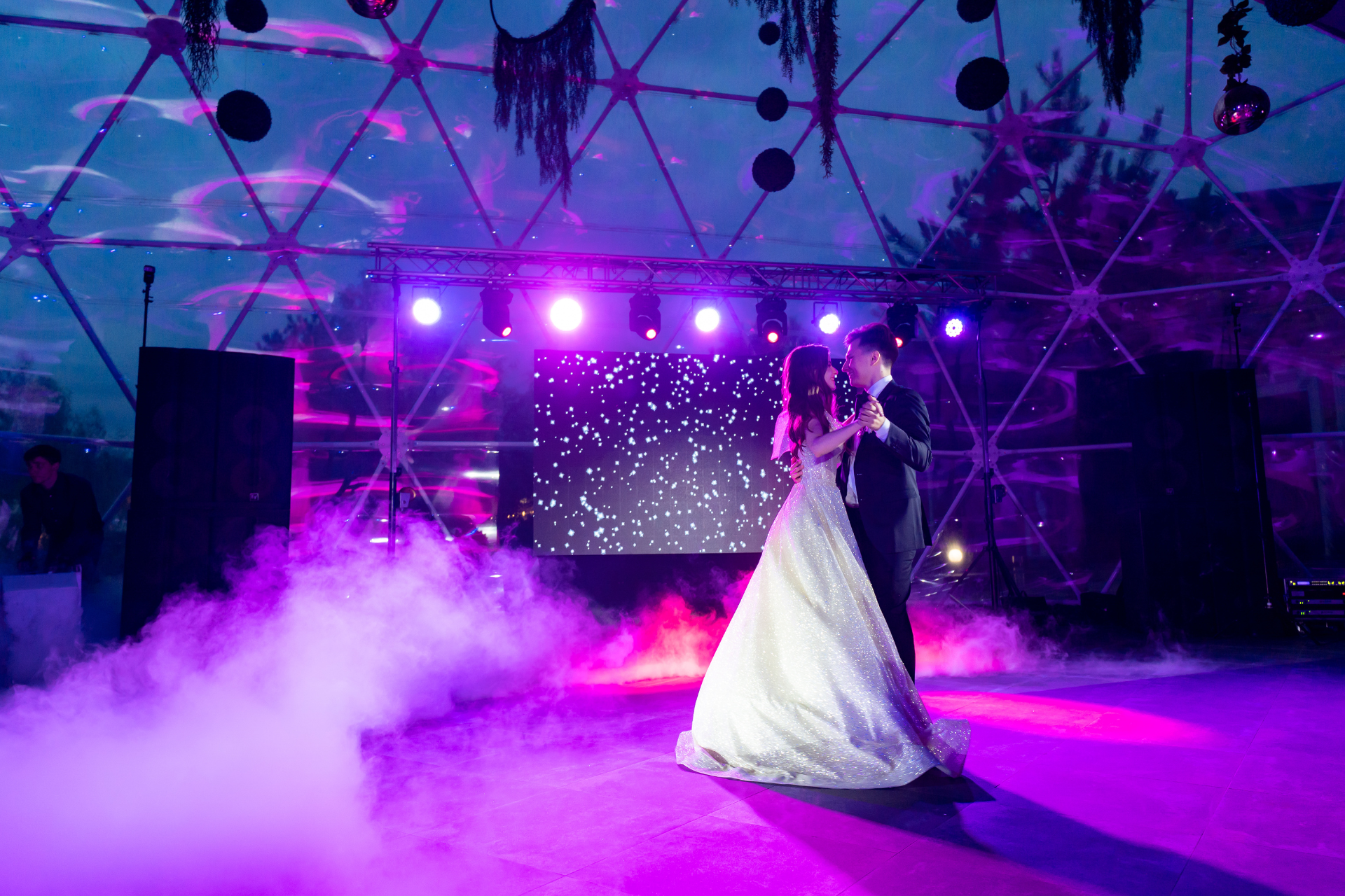 Bride and groom dancing at a wedding reception under purple lighting, with fog effects. The event is indoors.