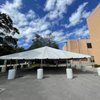 White tent over concrete, supported by cylindrical pillars, under a blue sky with clouds, next to a building.