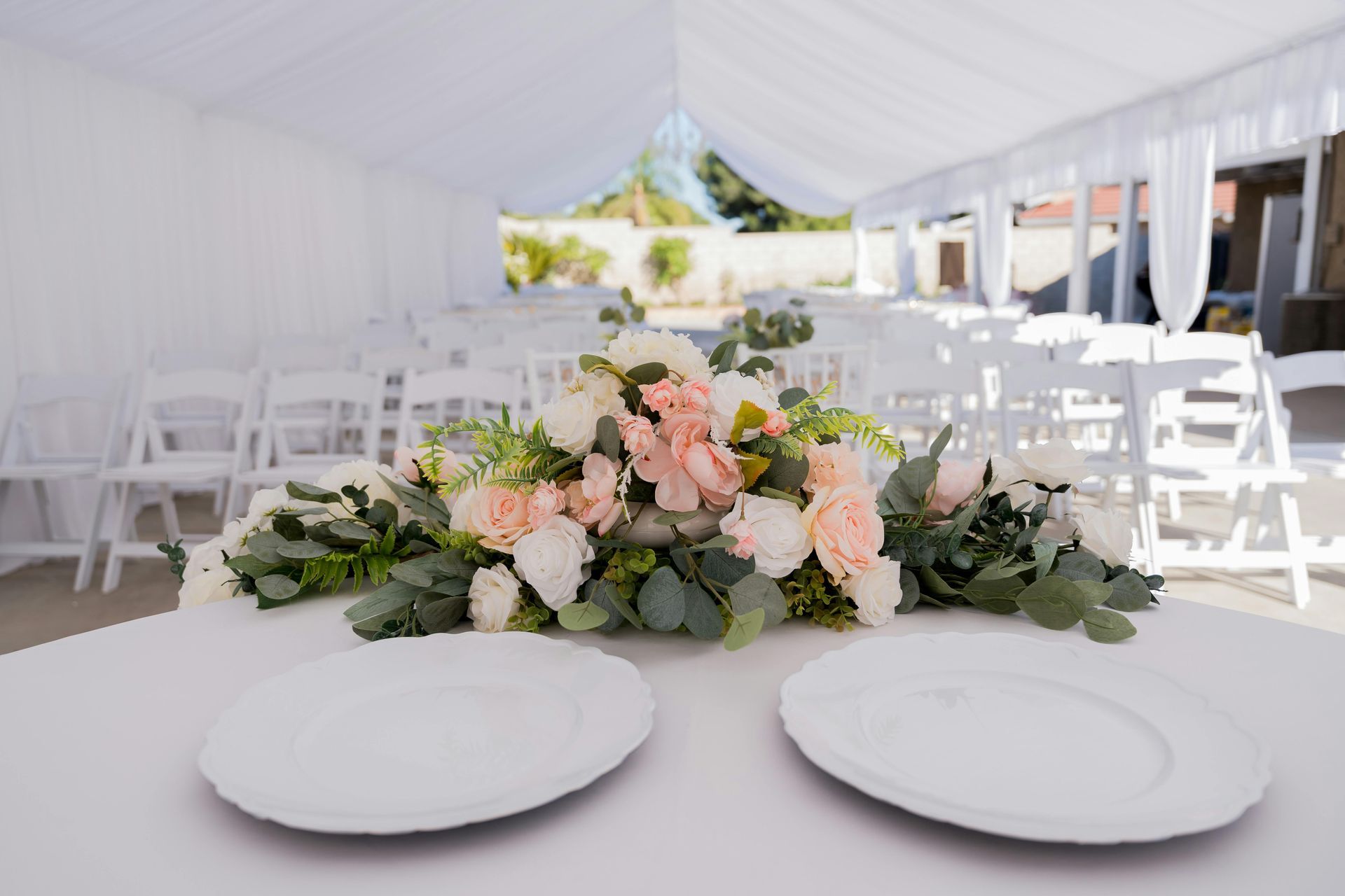 Wedding reception setup in a white tent on green grass. Long rows of chairs face a table with pink fabric, and round tables are set with floral centerpieces.