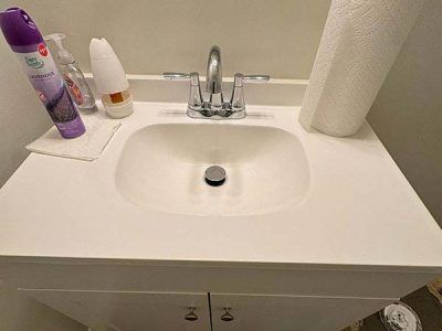 A white bathroom sink with cleaning supplies and paper towels on the countertop. A chrome faucet and drain are visible.
