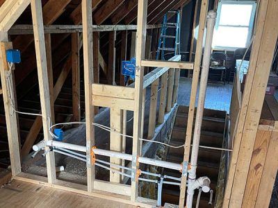 Interior view of unfinished construction in an attic. Framing with electrical boxes and plumbing pipes is installed near a staircase and window.
