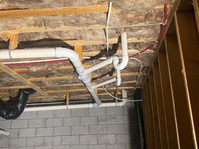White plumbing pipes snaking across a basement ceiling, connected to a drain and vent. Wood framing, insulation, and brick wall are visible.