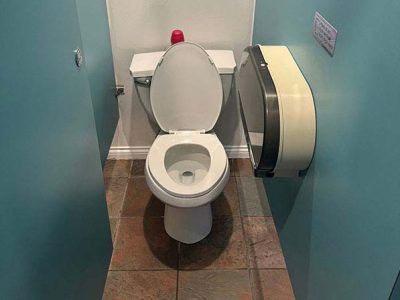 A toilet in a small bathroom with blue walls, a brown tiled floor, and a paper towel dispenser.