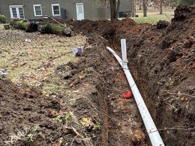 A trench in a yard holds white PVC pipes, dirt piled along the sides; a house is in the background.