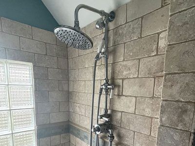 Shower with a large rain head and a secondary handheld showerhead, installed on a stone-tiled wall.