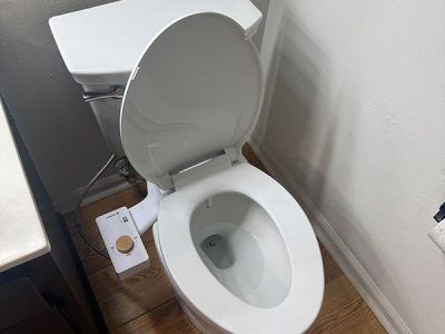 White toilet with the seat up, a bidet attachment, and a control panel are in a bathroom with light-colored walls and wood flooring.