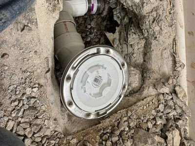 Close-up view of a plumbing cleanout in a concrete floor, with a gray fitting and white cap exposed in a hole.
