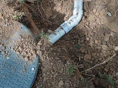 A section of buried PVC pipe emerging from the dirt next to a metal access cover. The ground is covered in loose soil and grass.